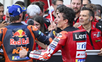 Two motorcycle racers in branded gear shake hands amid a cheering crowd, red Bull and Lenovo logos visible on their suits.