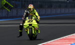 Motorcycle racer in neon yellow suit on a bright yellow bike (#43) performing a wheelie on a racetrack with a barrier in the background.