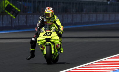 Motorcycle racer in neon yellow suit on a bright yellow bike (#43) performing a wheelie on a racetrack with a barrier in the background.