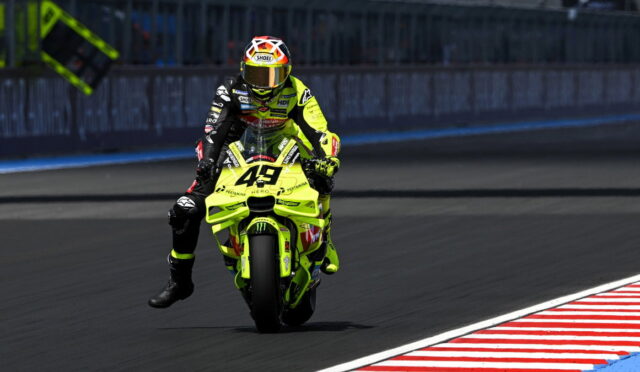 Motorcycle racer in neon yellow suit on a bright yellow bike (#43) performing a wheelie on a racetrack with a barrier in the background.