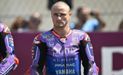 Male motorcycle racer in a purple Pramac Yamaha racing suit stands on the track, looking serious and focused.