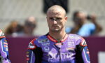 MotoGP rider in a purple Pramac racing suit stands on track, looking focused, with blurred spectators behind him.