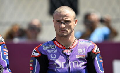 MotoGP rider in a purple Pramac racing suit stands on track, looking focused, with blurred spectators behind him.