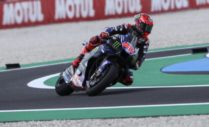 Motorcycle racer leaning into a sharp turn on a race track, wearing red and blue gear with sponsor decals on the bike.