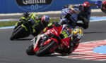 Motorcycle racers lean into a sharp turn on a race track, front rider in red with yellow helmet leading a pack.