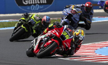 Motorcycle racers lean into a sharp turn on a race track, front rider in red with yellow helmet leading a pack.