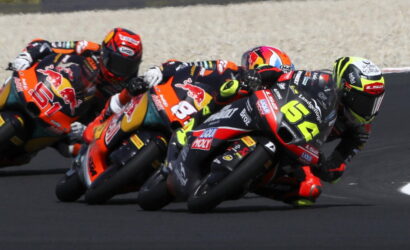 Motorcycle racers in bright racing suits lean into a tight left-hand turn on a smooth asphalt track, Biks numbered 65 and 84 visible in front.0So-phrase: racing scene with colorful bikes.