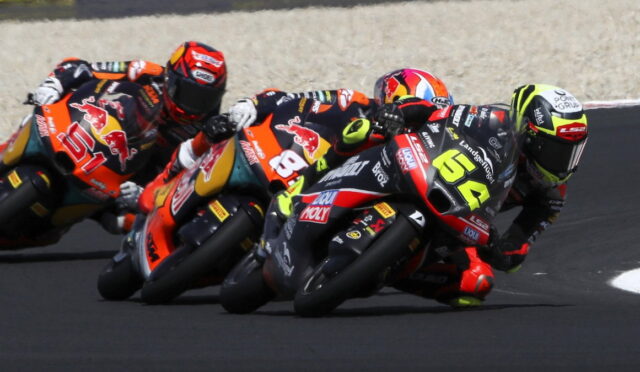 Motorcycle racers in bright racing suits lean into a tight left-hand turn on a smooth asphalt track, Biks numbered 65 and 84 visible in front.0So-phrase: racing scene with colorful bikes.