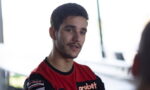 Portrait of a young man with short dark hair and light facial hair, wearing a red and black jersey with sponsor logos, looking slightly to the side.