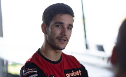 Portrait of a young man with short dark hair and light facial hair, wearing a red and black jersey with sponsor logos, looking slightly to the side.
