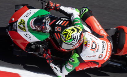 MotoGP rider in red, white, and green suit leaning into a sharp turn on a red Kawasaki race bike at speed.