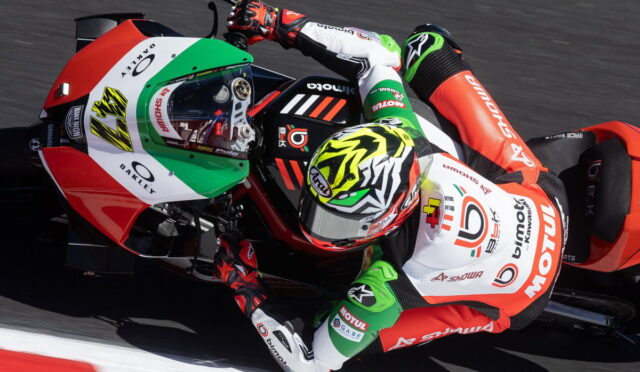 MotoGP rider in red, white, and green suit leaning into a sharp turn on a red Kawasaki race bike at speed.