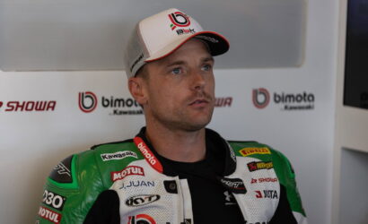 Moto racer in a green and white racing suit with sponsor logos, wearing a white cap, seated in a press area with Bimota/Kawasaki branding in the background.”