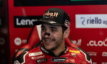 Portrait of a Ducati MotoGP rider smiling in a red racing suit and cap, in a sponsor-filled garage backdrop.