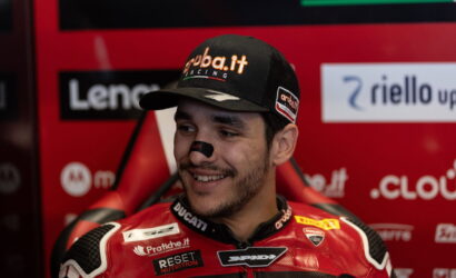 Portrait of a Ducati MotoGP rider smiling in a red racing suit and cap, in a sponsor-filled garage backdrop.
