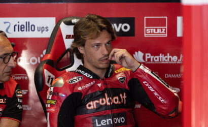 MotoGP rider in a red Ducati racing suit sits in a pit with sponsor banners behind, touching his lips thoughtfully.
