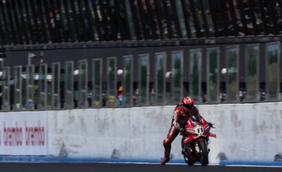 Rider in red racing suit leaning into a turn on a red motorcycle, number 11, on a racetrack with a concrete wall backdrop.