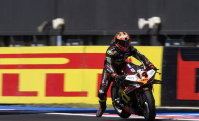 Racer in black and red suit on a sports motorcycle leaning into a turn on a racetrack, bike number 14 visible against yellow/red barriers.