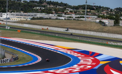Motorcycle racers on a curving race track with bright red, white, blue runoff painted around the bend and a hillside backdrop.