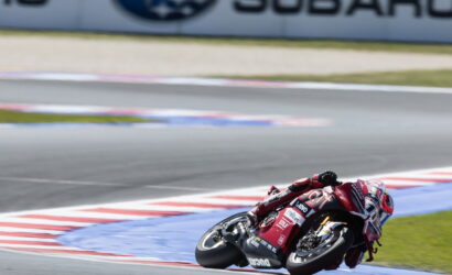 Motorcycle racer in red and white gear leans into a sharp turn on a race track, with a blurred Subaru banner in the background.