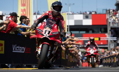 Moto racer in red Ducati bike #11 crossing the finish line as crowd cheers and banners fly by.