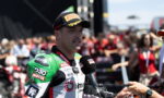 Motorcycle racer in green-and-white suit being interviewed with a handheld mic, crowd in the background at a race event, wearing a black Pirelli cap.