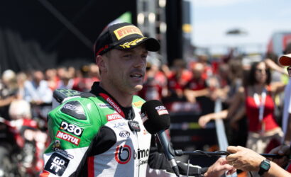Motorcycle racer in green-and-white suit being interviewed with a handheld mic, crowd in the background at a race event, wearing a black Pirelli cap.