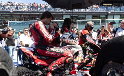Moto racer in red suit sits on a red sport bike under a large umbrella, surrounded by crew and spectators on race day.