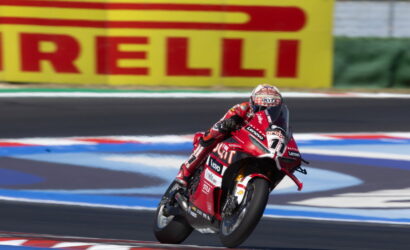 Racing motorcyclist in red suit leans into a turn on a track with a large Pirelli sign in the background.