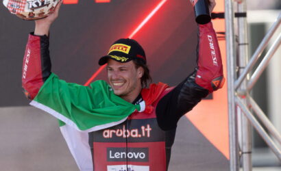 Race winner on a podium, arms raised in celebration with an Italian flag draped over shoulders, wearing a Pirelli cap and sponsor-labeled racing suit .