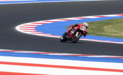 Motorcycle racer in a red racing suit leans into a sharp track corner on a dark asphalt circuit with blue and red striped runoff areas around the curve.