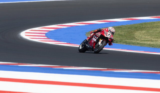 Motorcycle racer in a red racing suit leans into a sharp track corner on a dark asphalt circuit with blue and red striped runoff areas around the curve.