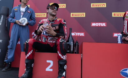 Racer in red suit seated on 2nd place podium, holding gifts, with sponsor backdrop and teammate in blue jumpsuit beside him.