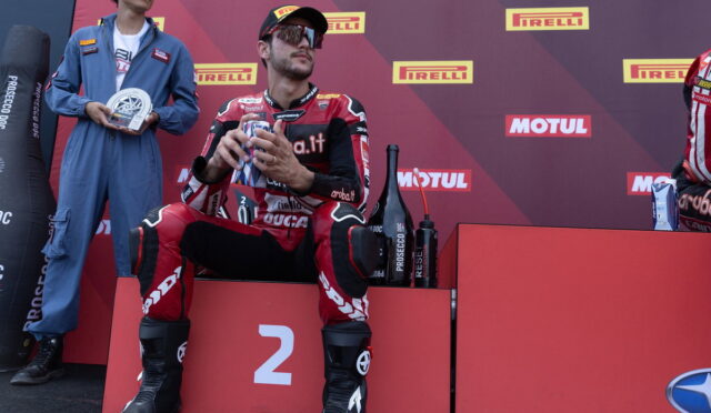 Racer in red suit seated on 2nd place podium, holding gifts, with sponsor backdrop and teammate in blue jumpsuit beside him.
