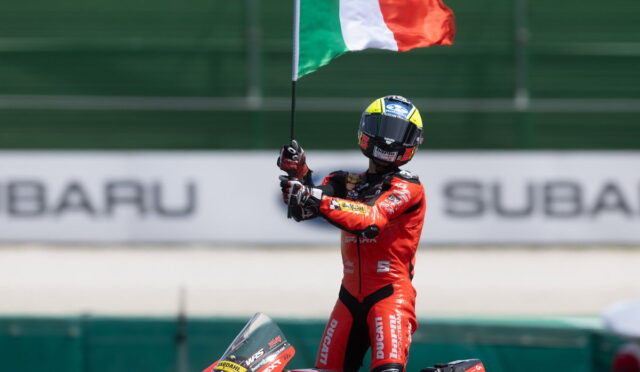 Motorcycle racer in a red Ducati suit raises an Italian flag on a race track beside the bike.
