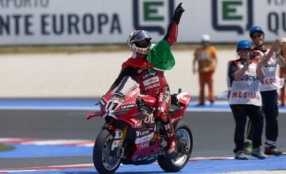 Rider in red Ducati gear on bike #17 celebrates, raising one arm as they crest a blue race track with spectators in the background.