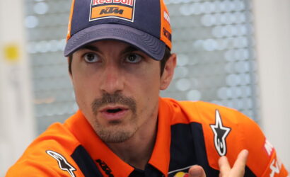 Close-up of a man in an orange KTM/Red Bull racing cap and matching racing jersey, looking to the side as if talking or listening.