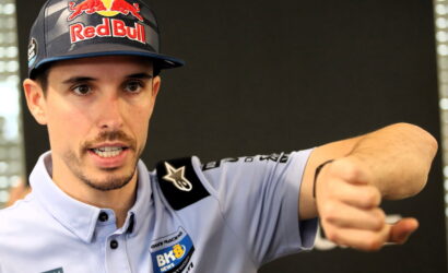Portrait of a man wearing a Red Bull cap and light blue racing shirt, extending his fist toward the camera.