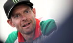 Close-up of a man wearing a black cap with a green Monster logo, looking surprised or curious, wearing a green and white shirt with a red collar.