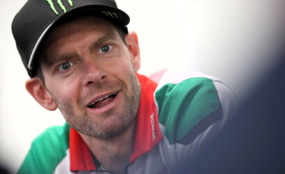 Close-up of a man wearing a black cap with a green Monster logo, looking surprised or curious, wearing a green and white shirt with a red collar.