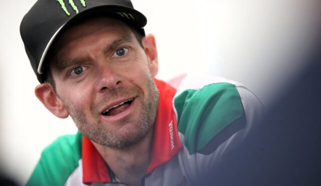 Close-up of a man wearing a black cap with a green Monster logo, looking surprised or curious, wearing a green and white shirt with a red collar.