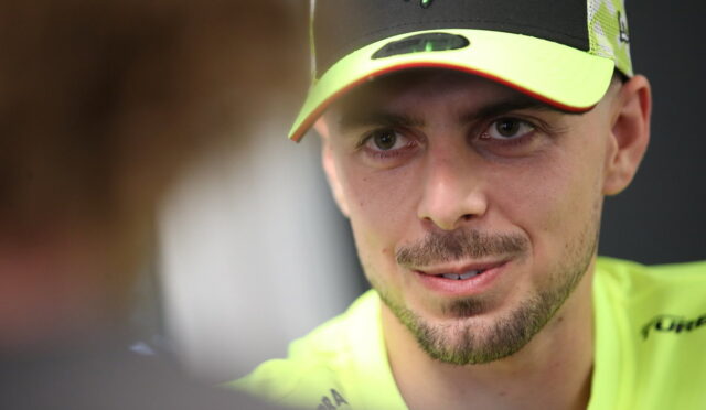 Close-up of a man wearing a neon yellow cap and shirt, looking at someone off-camera while speaking.