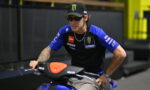 Rider in a blue and black Yamaha sponsorship shirt sits on a blue motorcycle in a pit area.