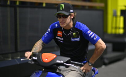 Rider in a blue and black Yamaha sponsorship shirt sits on a blue motorcycle in a pit area.