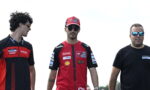 Three men stand outdoors in Ducati racing team gear; two wear red-and-black shirts and caps, the third in a black T-shirt with sunglasses.