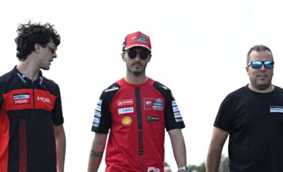 Three men stand outdoors in Ducati racing team gear; two wear red-and-black shirts and caps, the third in a black T-shirt with sunglasses.