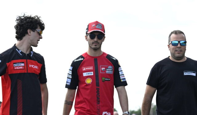 Three men stand outdoors in Ducati racing team gear; two wear red-and-black shirts and caps, the third in a black T-shirt with sunglasses.