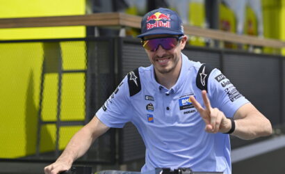 Rider in a light-blue polo with sponsor patches, wearing a Red Bull cap and mirrored sunglasses, flashing a peace sign beside his bike