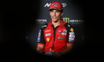 Male motorcycle racer in a red Ducati team polo and cap, speaking at a press event with Monster Energy branding behind him.