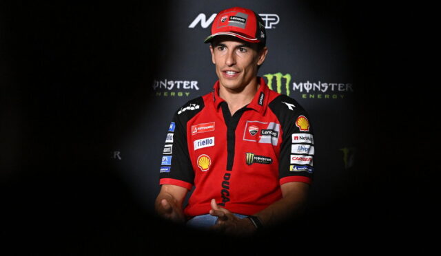 Male motorcycle racer in a red Ducati team polo and cap, speaking at a press event with Monster Energy branding behind him.